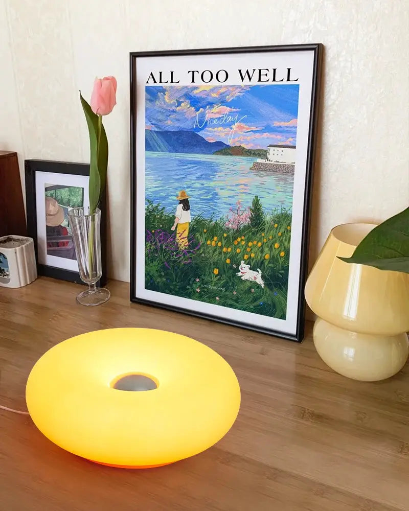 Bauhaus donut-shaped USB-powered table lamp in warm yellow by Simples Market on a wooden floor beside framed art