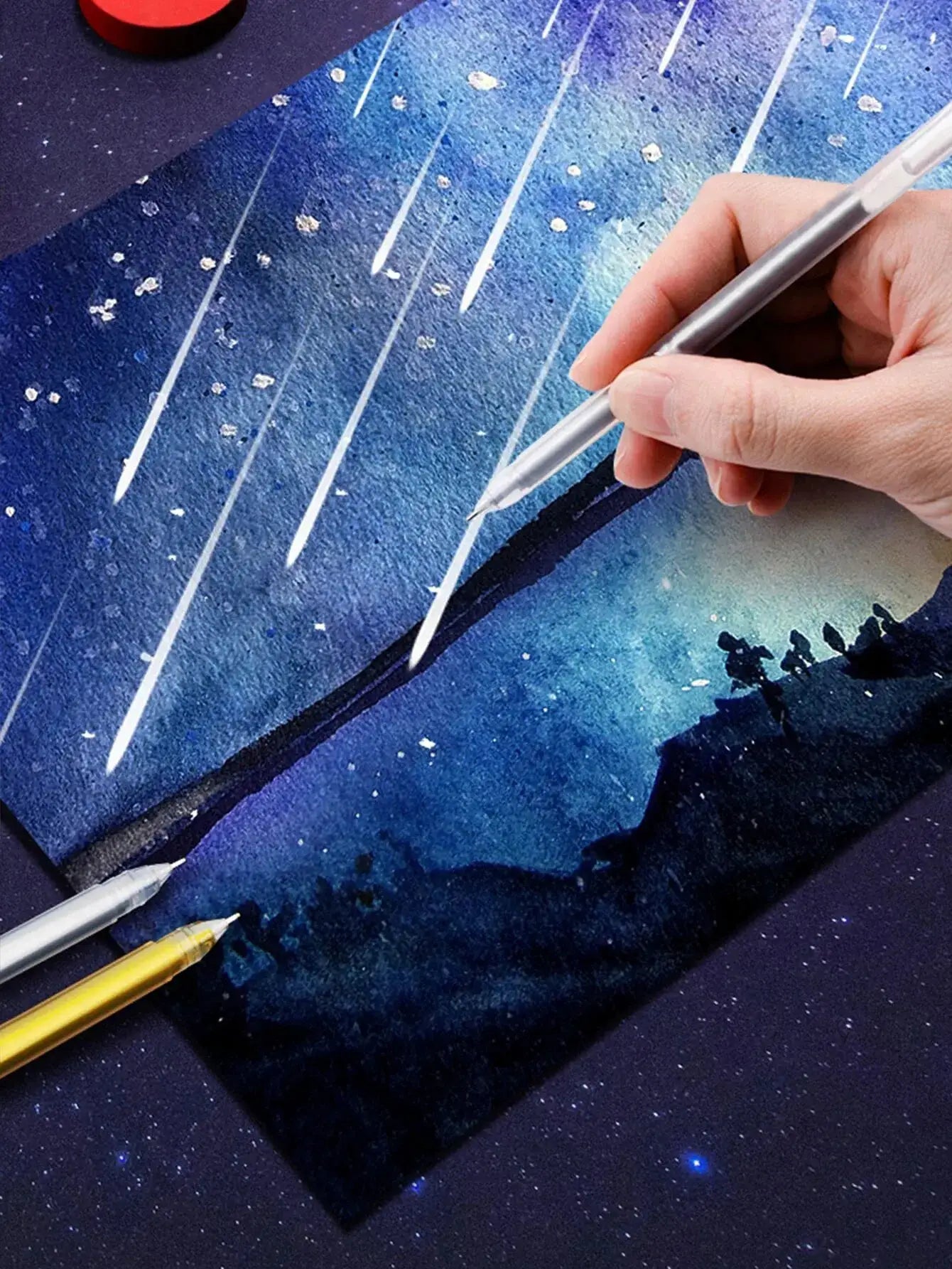 12-color gel pen set with silver barrel held by hand over blue artwork; 0.5mm fine tip