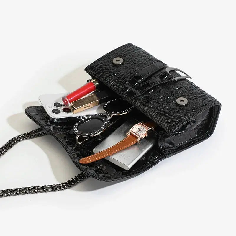 Simples Market black croc-embossed crossbody bag with chain strap; phone, lipstick, sunglasses and watch visible.