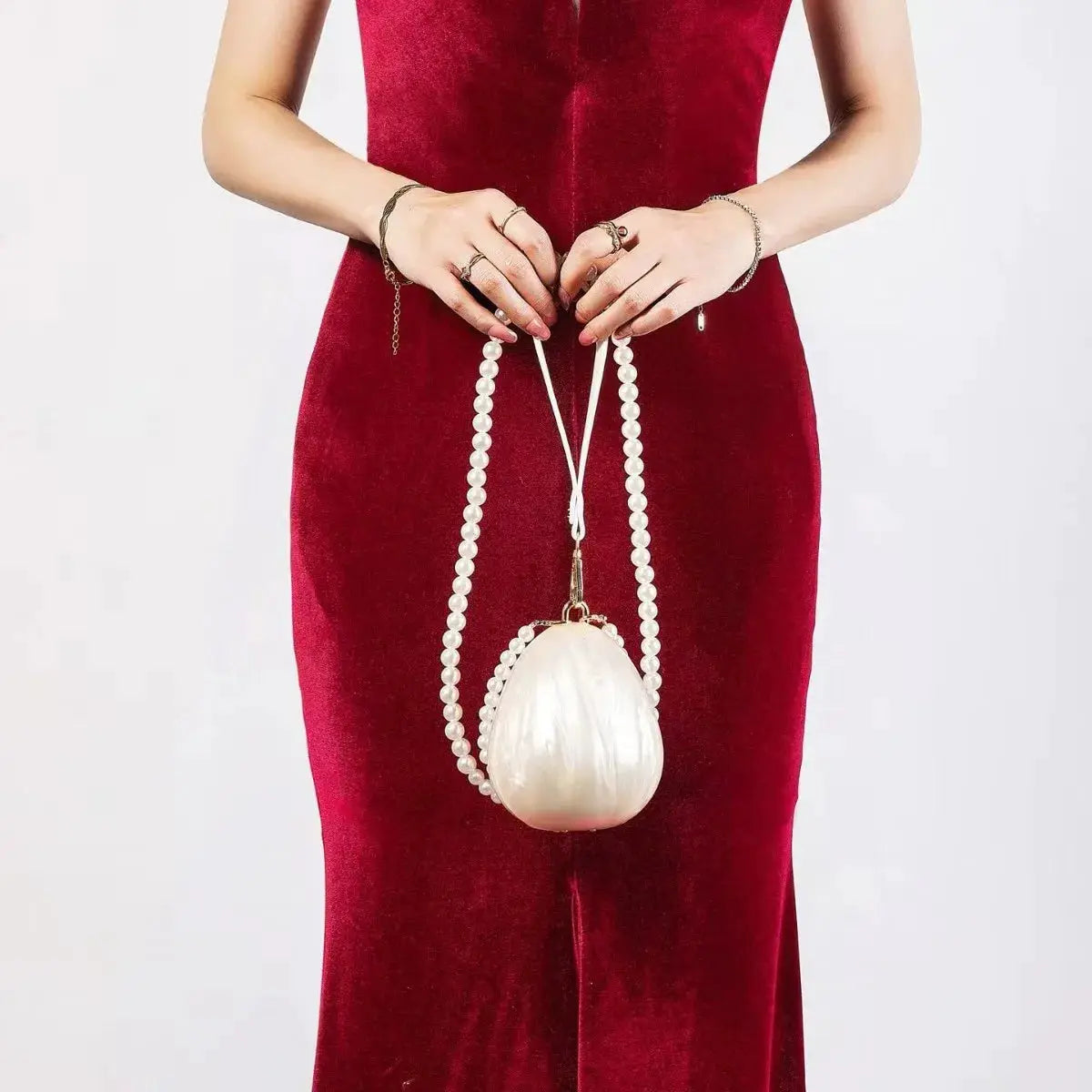 Egg-shaped pearl beaded clutch bag with a white shell-like body and pearl strap, held by a model in a red dress.