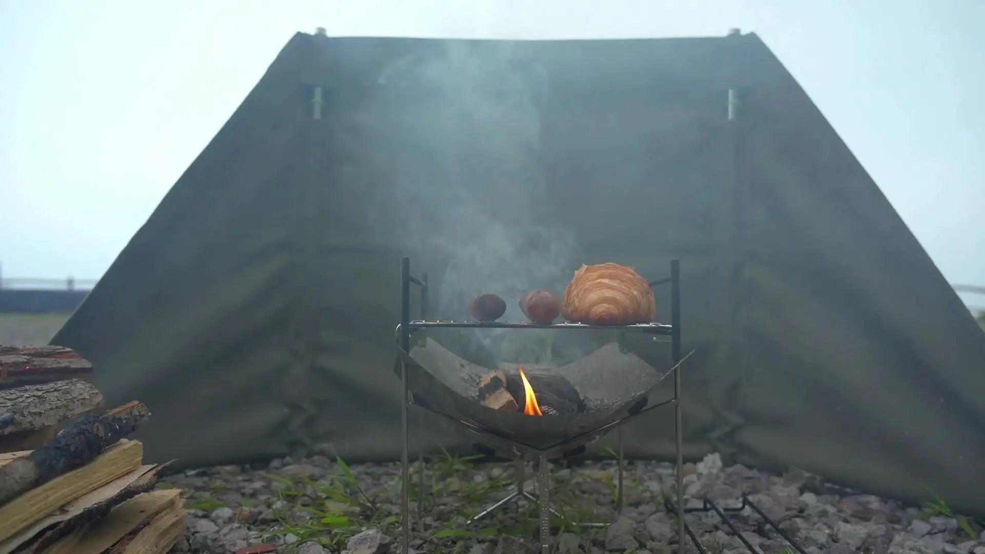 Stainless steel portable folding BBQ grill for camping; bread rolls on the grate near a smoky campfire.