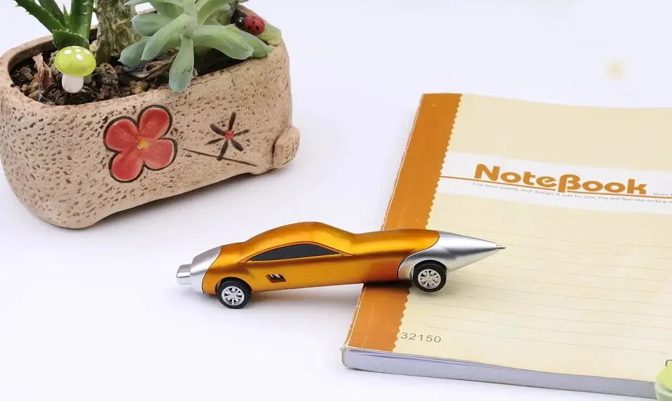 Car-shaped ballpoint pen in metallic orange, candy-color finish, on a notebook beside a potted plant.
