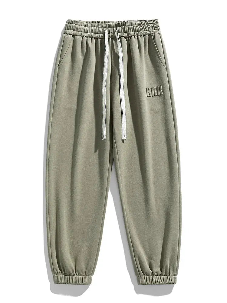 Olive green cotton/polyester jogger pants with elastic waist, white drawstrings, cuffed ankles, loose fit, thigh patch.