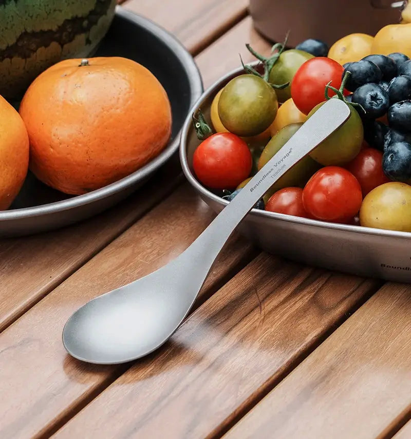 Boundless Voyage titanium spoon in silver, Simples Market outdoor flatware, on a wood table beside tomatoes