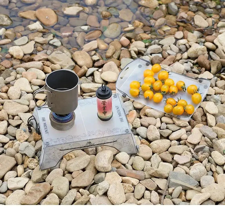 Ultralight folding mini table gray PP on rocky ground with cooking gear; tray of yellow fruits.