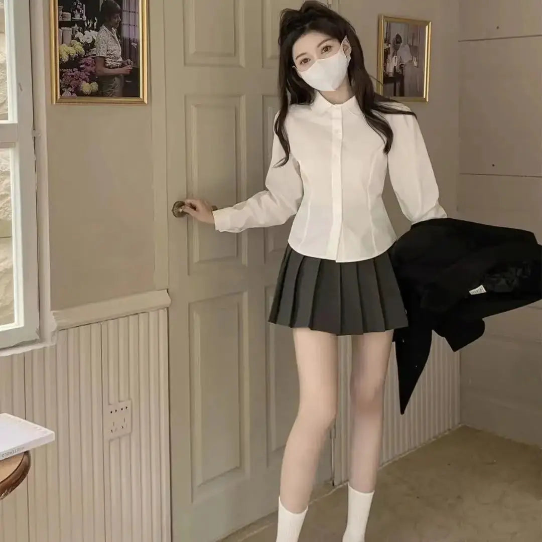 White women's button-down shirt, slim long sleeves, shown with a black pleated mini skirt for a preppy school look.
