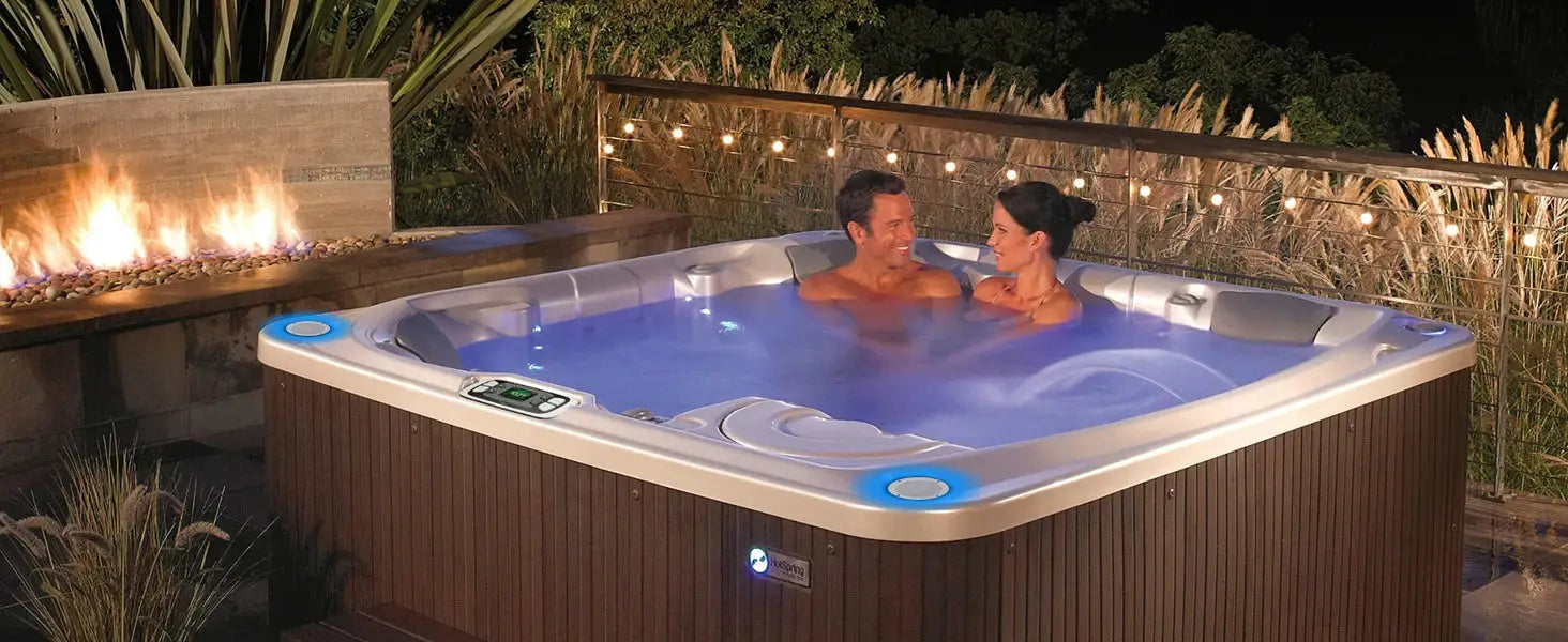 Hot tub with wooden exterior and blue water; two people enjoy a night soak under string lights.