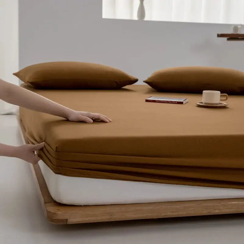 Soft cotton fitted sheet in brown with elastic edge on bed, two pillows visible