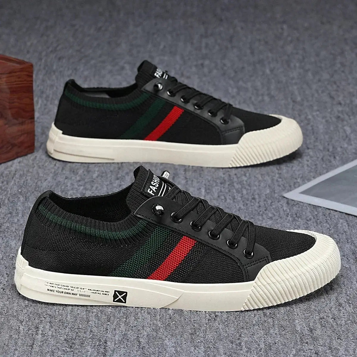 Two black lace-up sneakers with red and green stripes, white rubber toe caps, and chunky white soles on a gray carpet.