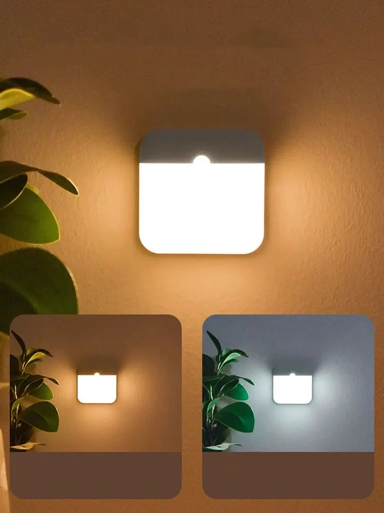 Motion sensor LED square wall light, white with a gray top, mounted on a wall beside a plant, emitting a warm glow