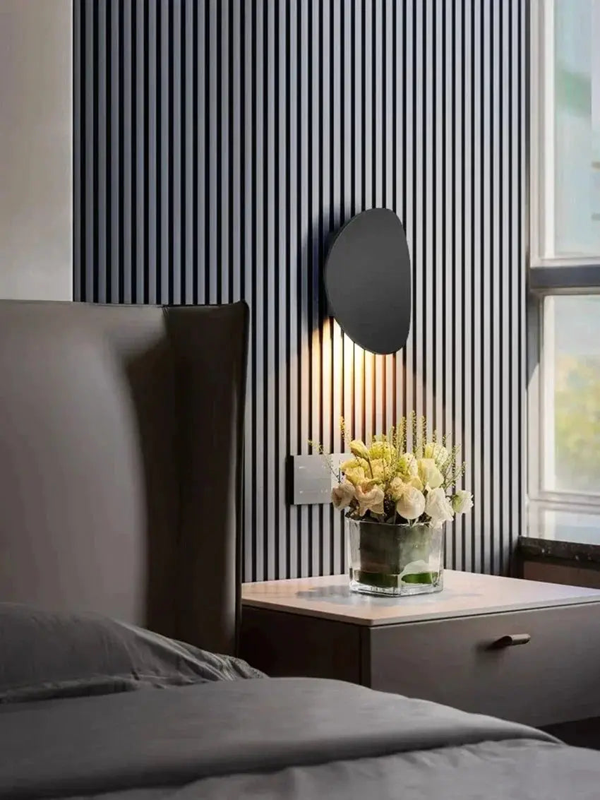 Simples Market outdoor LED wall lamp; round black wall sconce casting warm light beside the bed on striped wall.