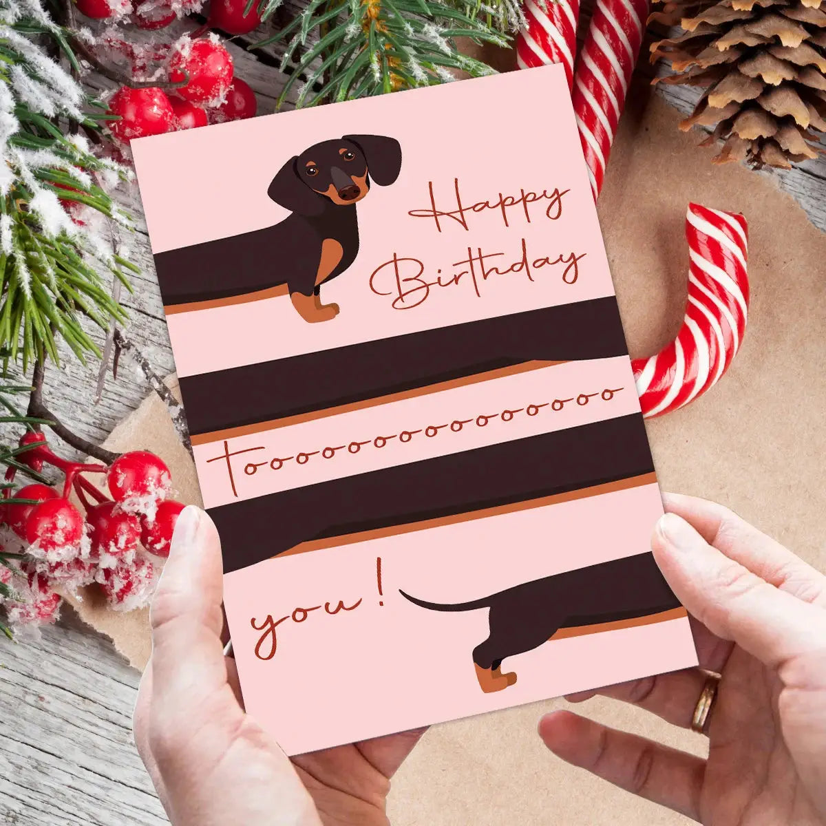 Whimsical dachshund birthday greeting card in pink with brown stripe accents, held by hands