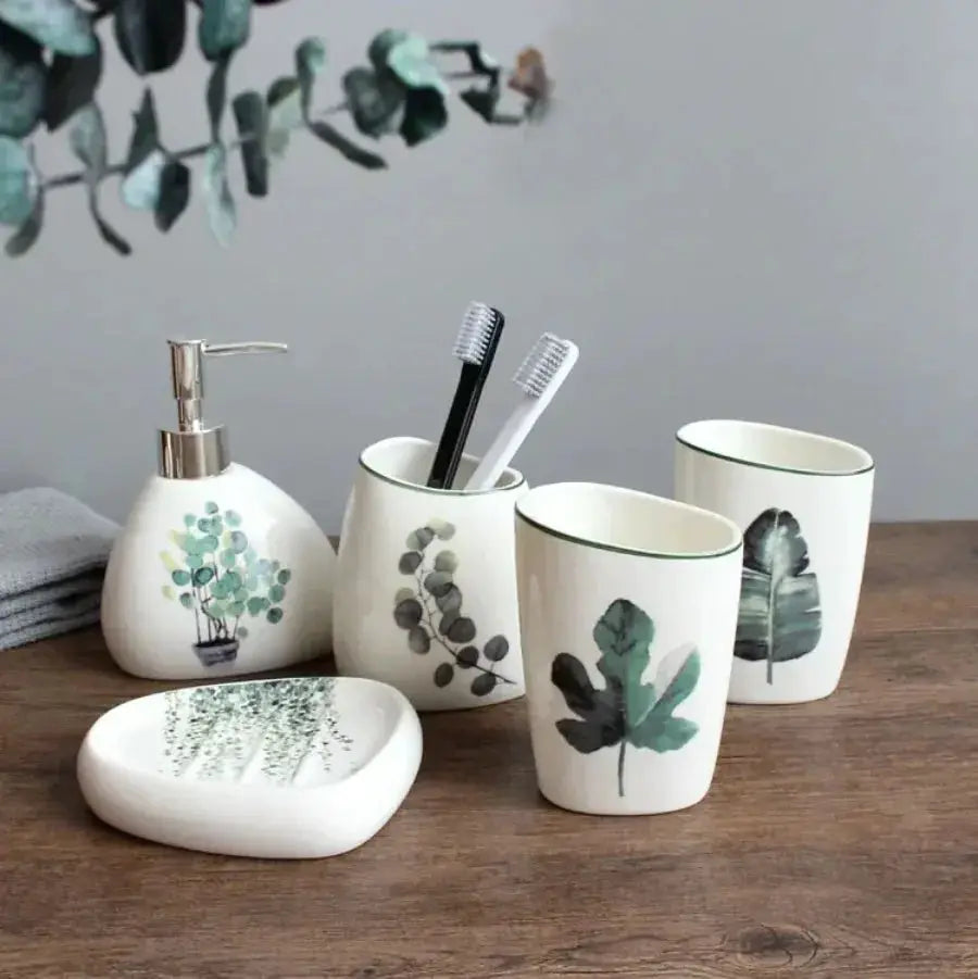 White ceramic bathroom accessory set with green botanical prints; includes soap dispenser, toothbrush holder, cups, and soap dish.