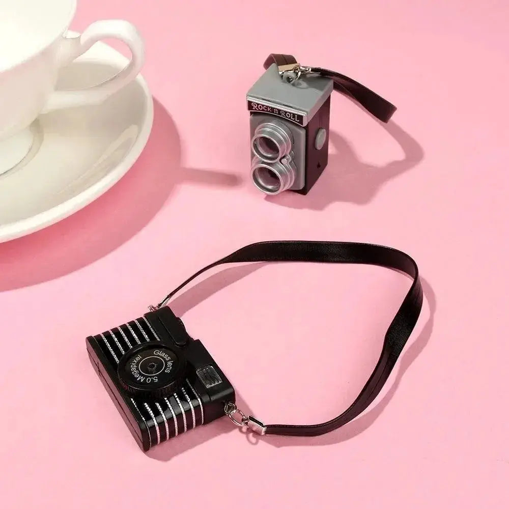 Two dollhouse miniature cameras: a black retro camera with strap and a silver twin-lens mini camera on a pink background.