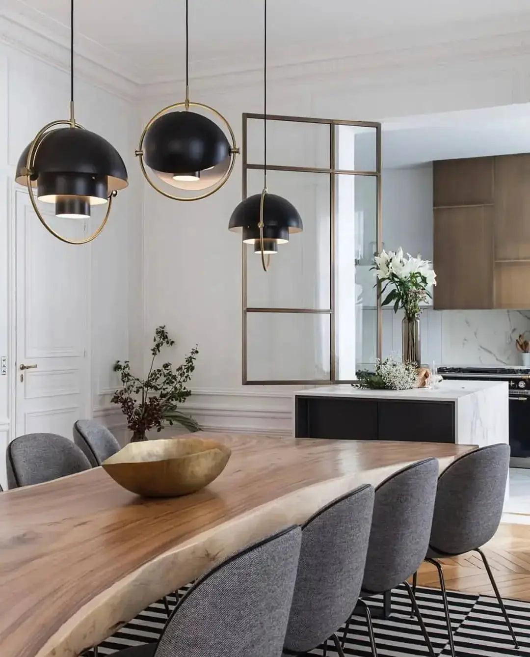 Pendant lights, three black iron domes with gold rings, hanging over a wooden dining table in a modern kitchen.