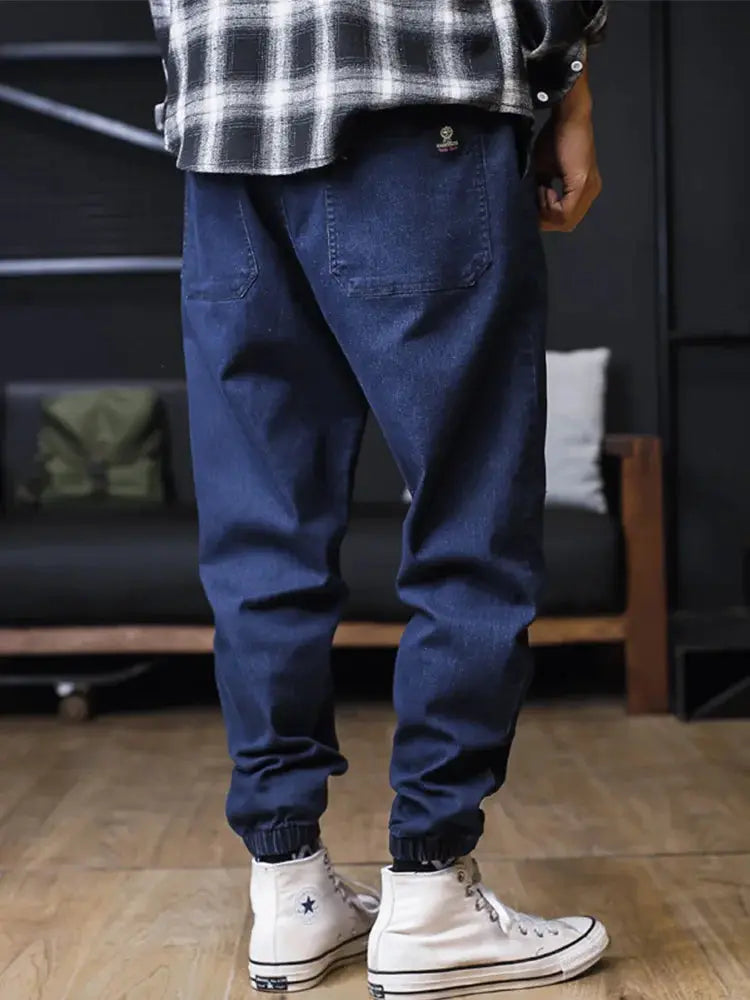 Plus-size men's denim jogger jeans by Simples Market, loose harem-style cargo pants with ankle cuffs in dark indigo.