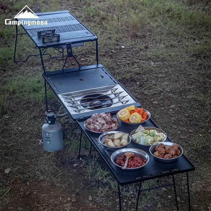 CampingMoon CK-25BK-1PIGT black metal camping table with stove top, six bowls of food, and propane canister.