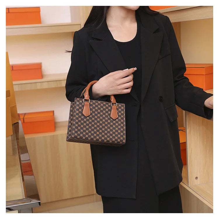 Small square top-handle handbag in plaid PU leather with tan handles, held by a woman in a black blazer