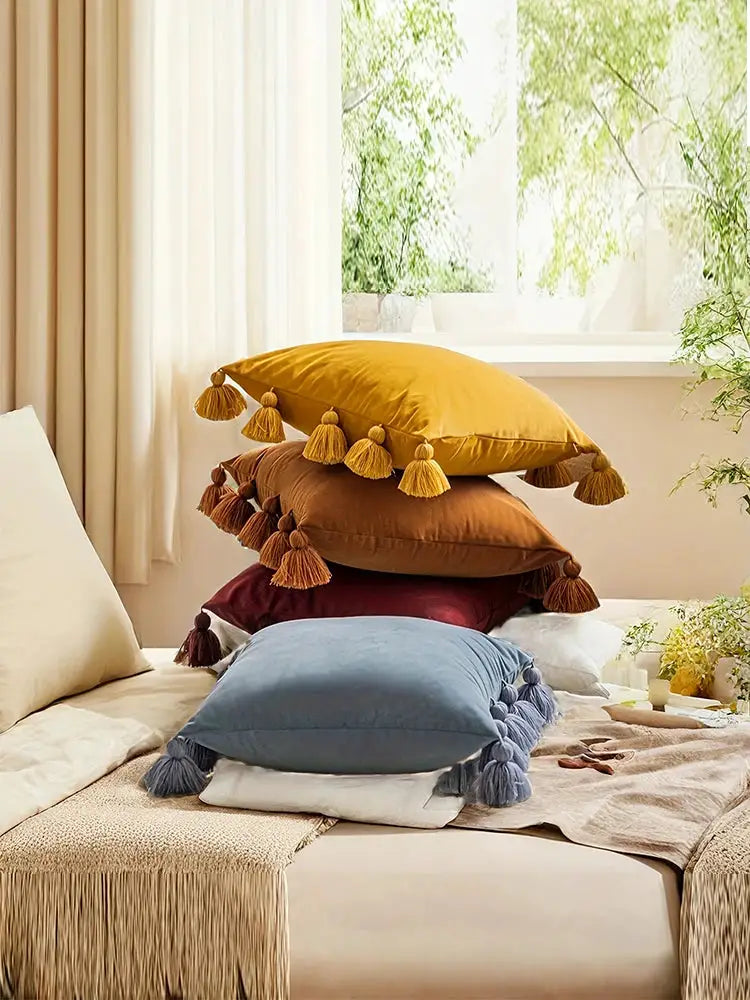 Decorative tassel throw pillow set stacked on bed in yellow, tan, burgundy, and blue