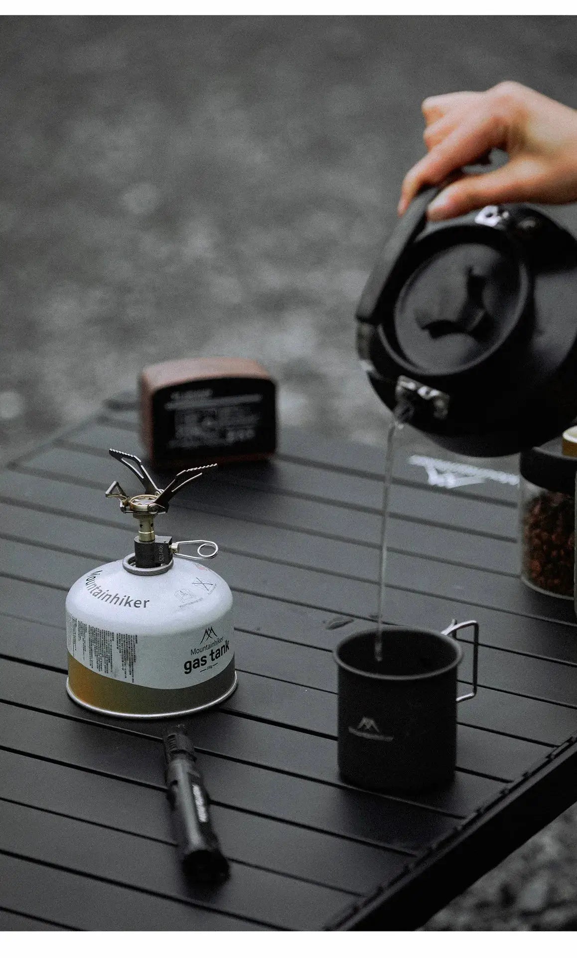 MK-3000W mini camping gas burner on a 48g canister with folding arms on a black camping table beside a kettle and mug
