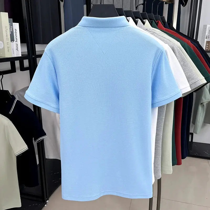 Light blue cotton men's polo shirt on a hanger, with other colored polos on a rack behind.