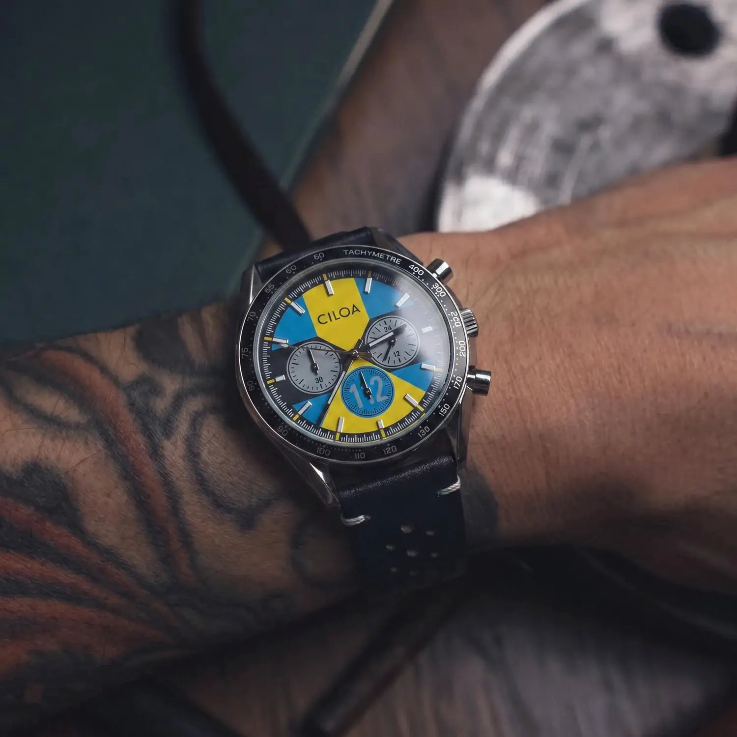 CILOA men's racing chronograph watch with vintage blue and yellow dial, tachymeter bezel, black perforated leather strap.