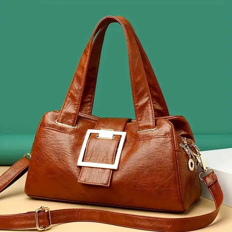 Top-handle leather handbag in brown with a large gold square buckle and detachable shoulder strap.