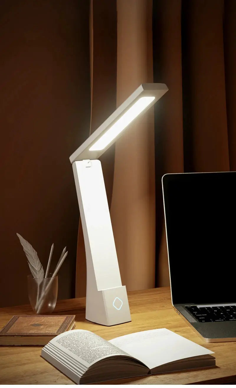 LED desk lamp by Simples Market, white foldable arm with built-in LED bar on a wooden desk beside a laptop