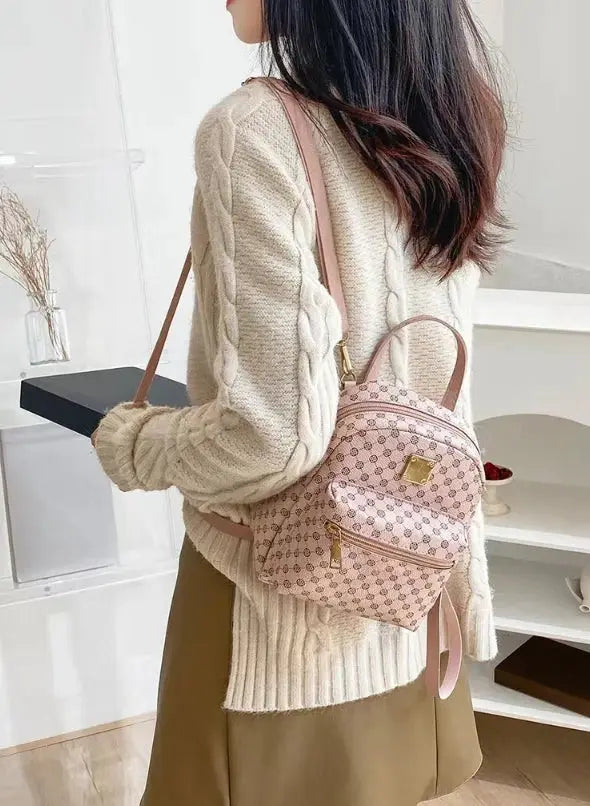 Backpack Simples Market, pink patterned with gold hardware and top handle, worn by a woman in a cream sweater.