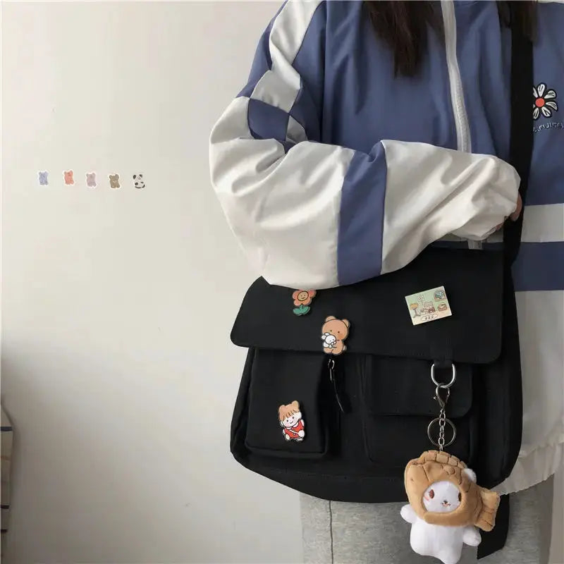 Women's black canvas messenger bag with flap, front pockets, decorative patches, and a plush keychain