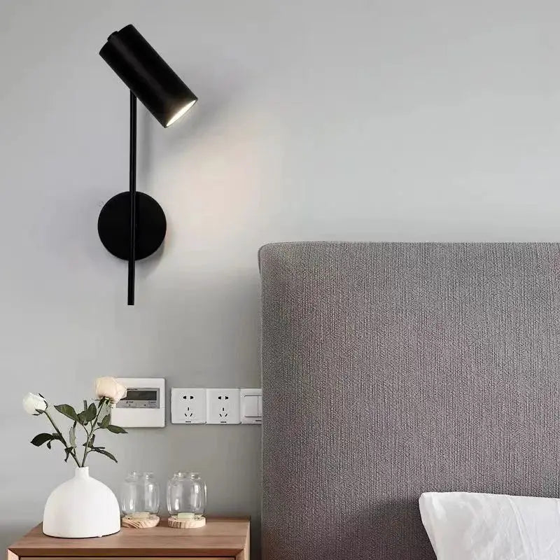 Rotatable black wall lamp with iron frame and cylindrical plastic shade, hotel-style bedside sconce.