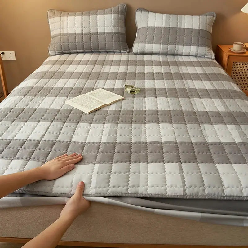 Quilted plaid mattress protector on a bed with 2 grey and white pillows; breathable, antibacterial, comfortable.