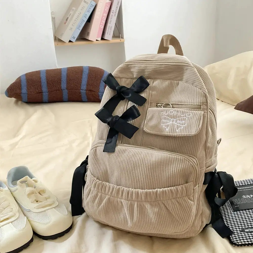 Beige corduroy backpack with black ribbon bows, front zip pocket, and elastic side pocket