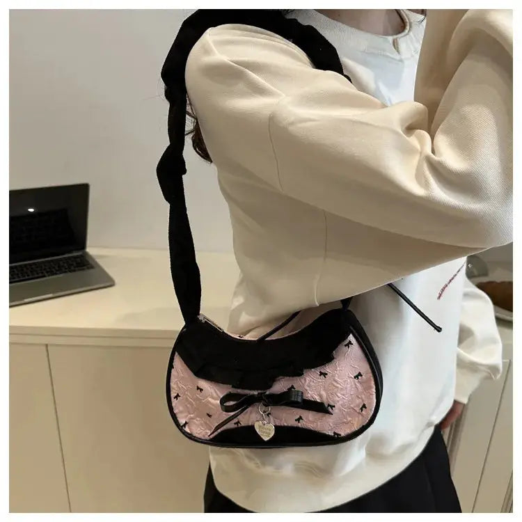 Crescent pink handbag with black trim, bow, and heart pendant; compact shoulder bag.
