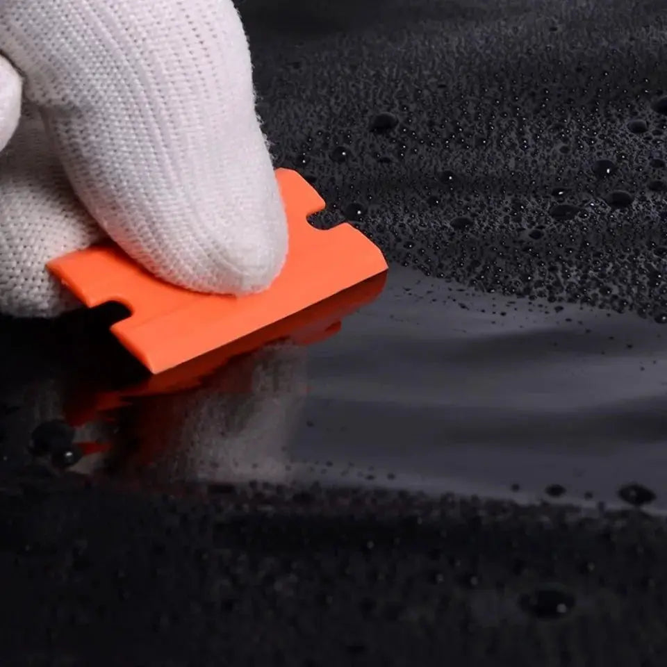 Orange plastic razor blade scraper used on glass by a gloved hand.