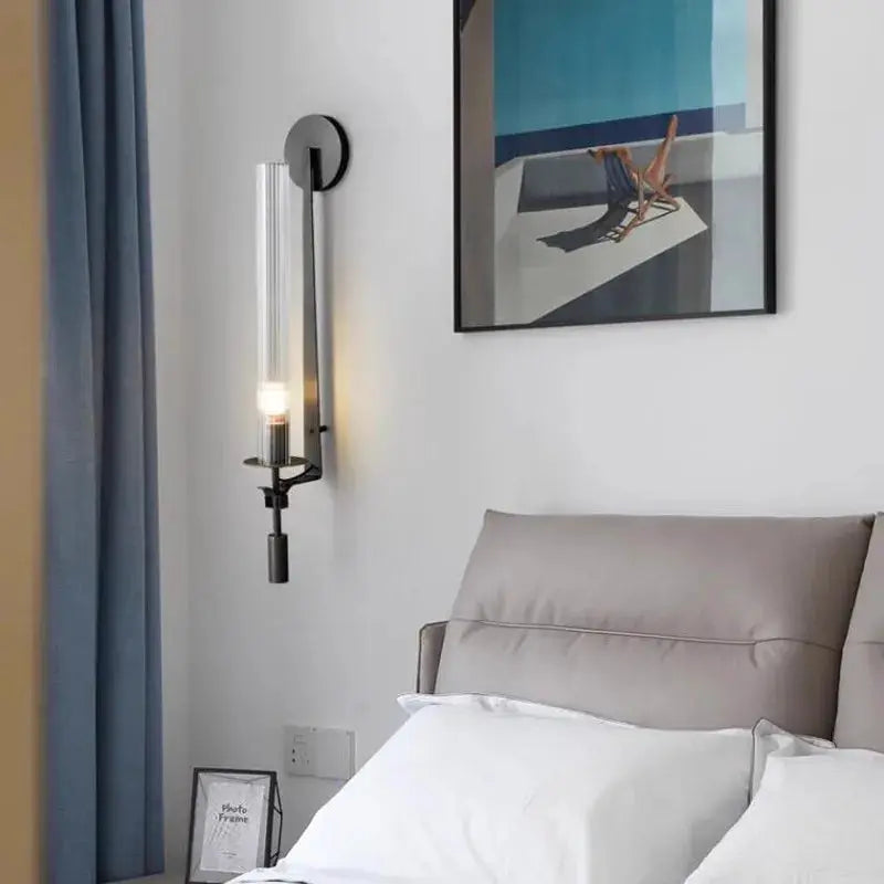 Modern copper wall sconce lamp with a clear glass tube, mounted on a bedroom wall beside the bed.