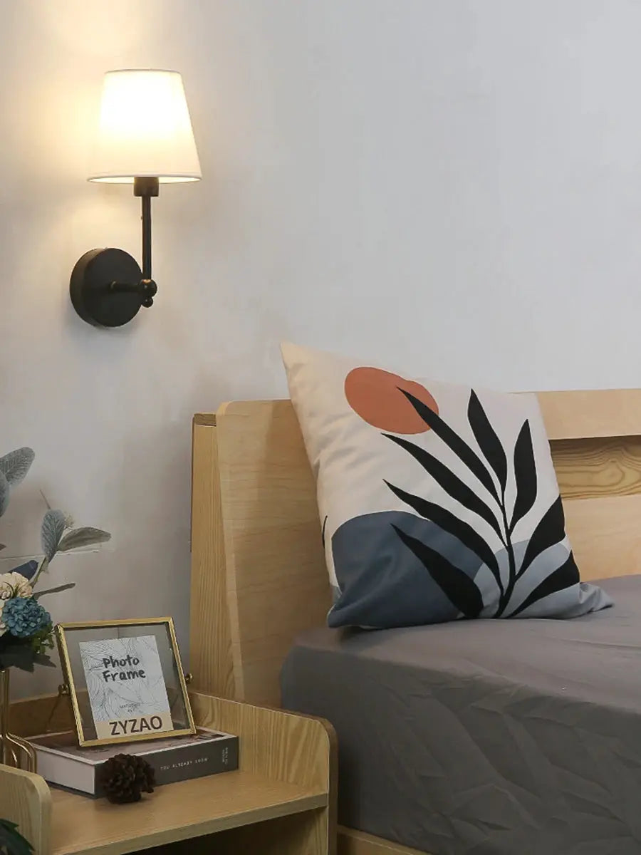 Wall lamp with black metal base and white fabric shade, mounted beside a wooden bed.
