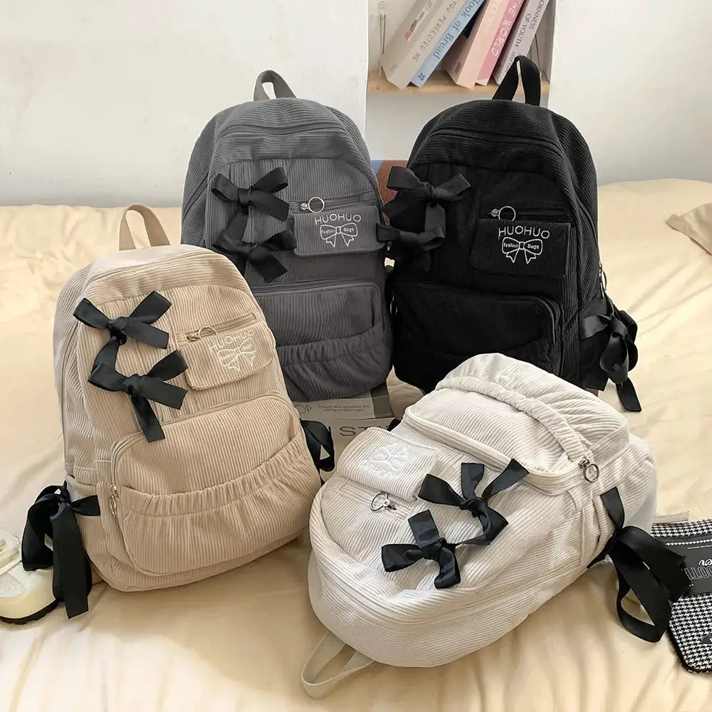 Backpacks set of four by Simples Market in beige, gray, black, and cream with corduroy fabric and bow accents.