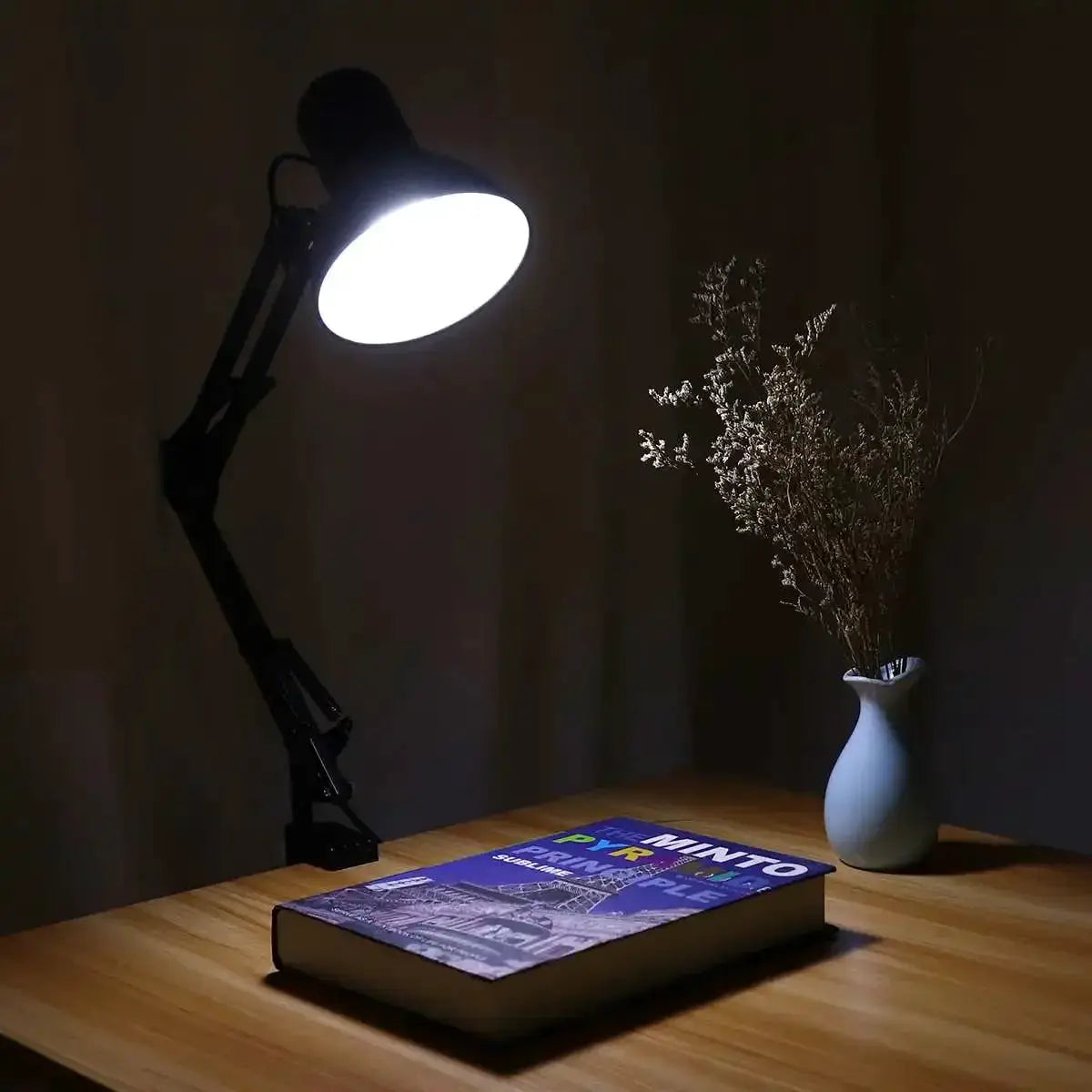 Simples Market Swing Arm Clamp Desk Lamp, black metal, adjustable, lit over a wooden desk with a book and vase.