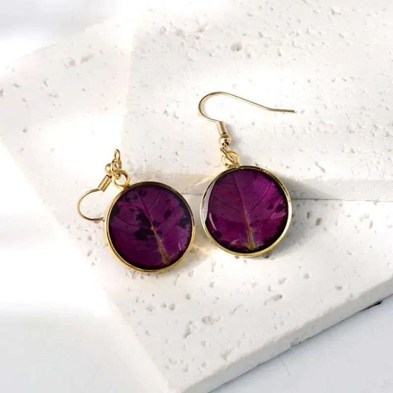 Pressed flower earrings pair by Simples Market, purple epoxy resin in gold circular frames.
