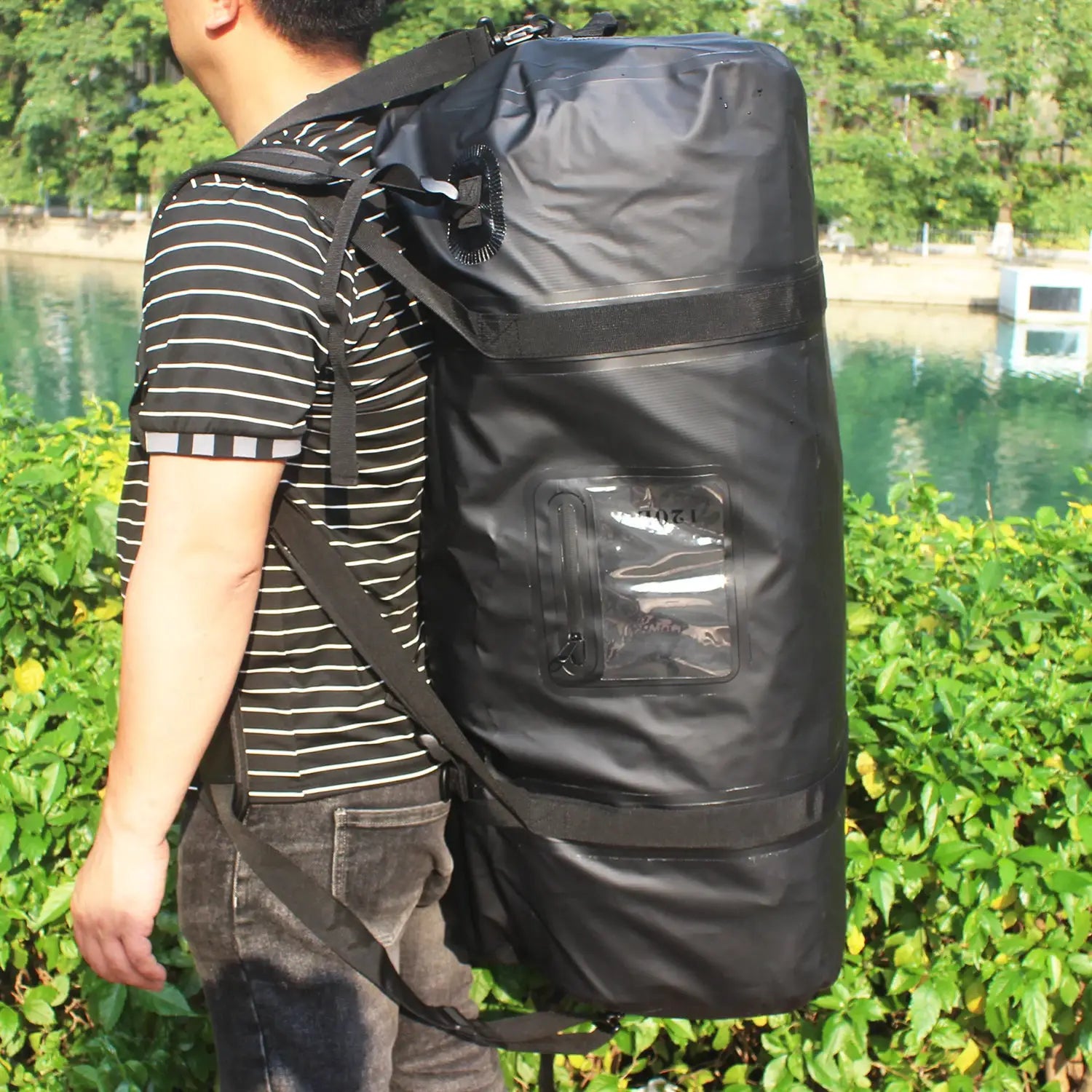 Black waterproof duffle backpack (40–120L) with clear side pocket worn by a person outdoors.