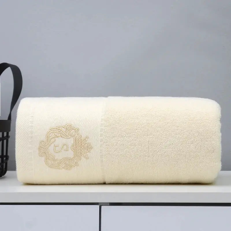 White cotton towel set by Simples Market, beige embroidered crest, folded on a bathroom counter.