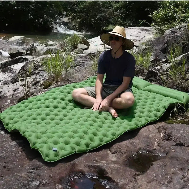 Double air mattress camping bed, green inflatable pad on a rocky riverbank with a seated person