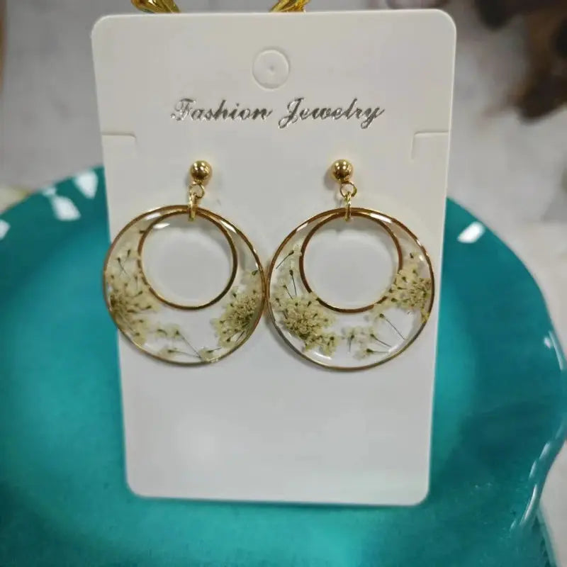 Gold-tone hoop earrings with clear resin encasing pressed white flowers, geometric double-hoop design.