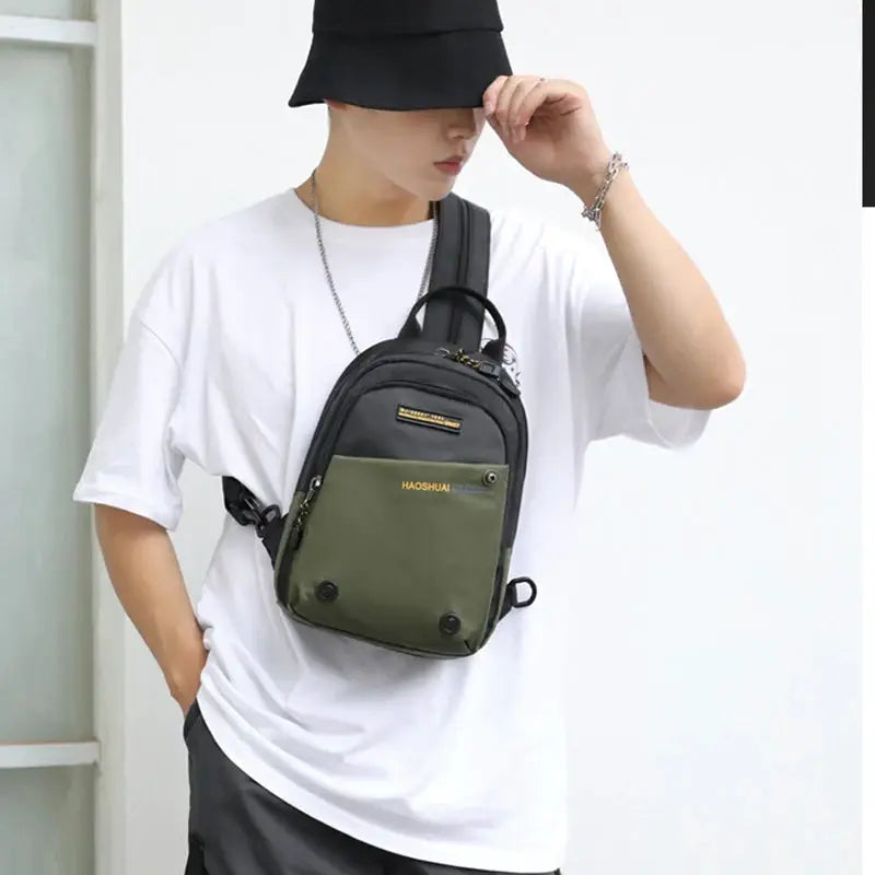 Men's small nylon backpack with black body and olive green front pocket featuring gold-tone zippers, crossbody strap