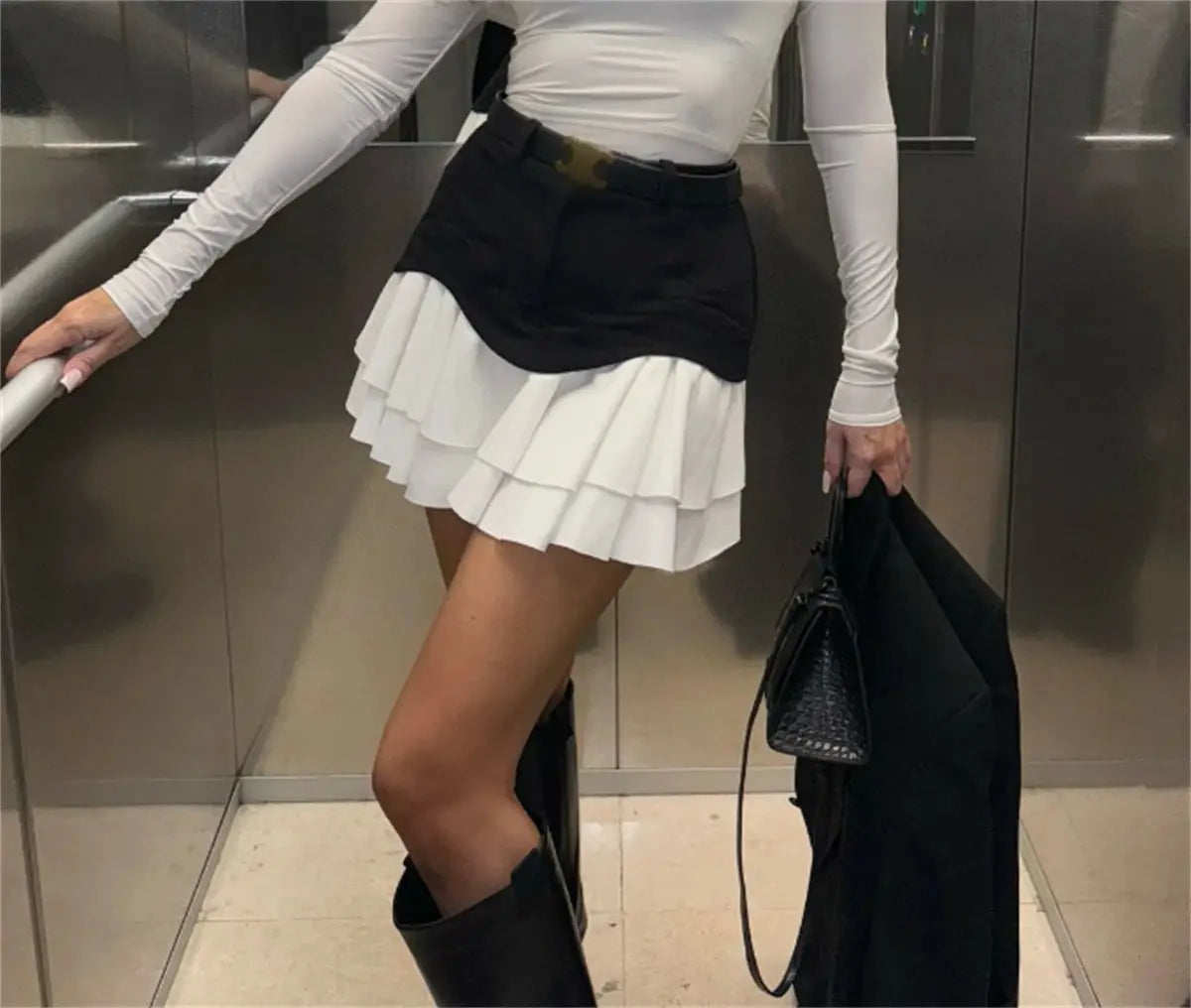 Chic Ruffles white pleated mini skirt with black waist overlay; white top, black boots, carrying a black jacket
