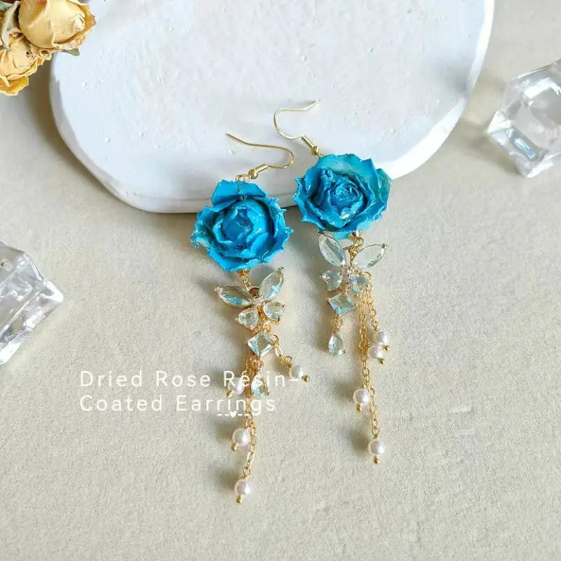Long earrings featuring blue epoxy resin rose with gold-tone chain, crystal leaf charms and pearl drops