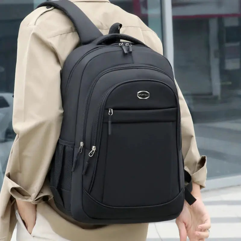 Backpack, black, large-capacity with multiple zip pockets, worn by a person in a beige coat.