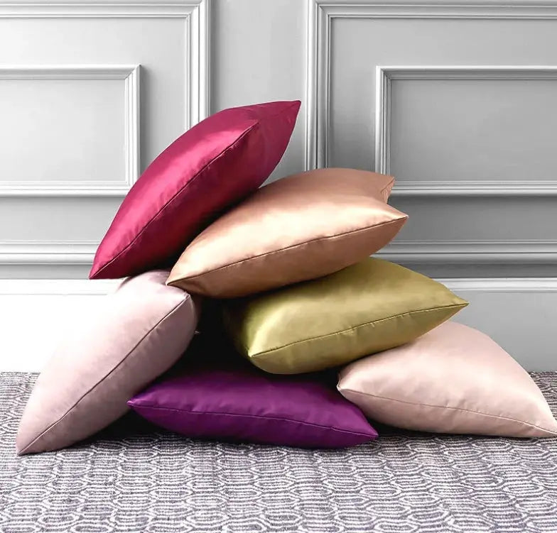 Six satin pillow covers in magenta, peach, olive, pink, purple and beige stacked on a patterned rug.
