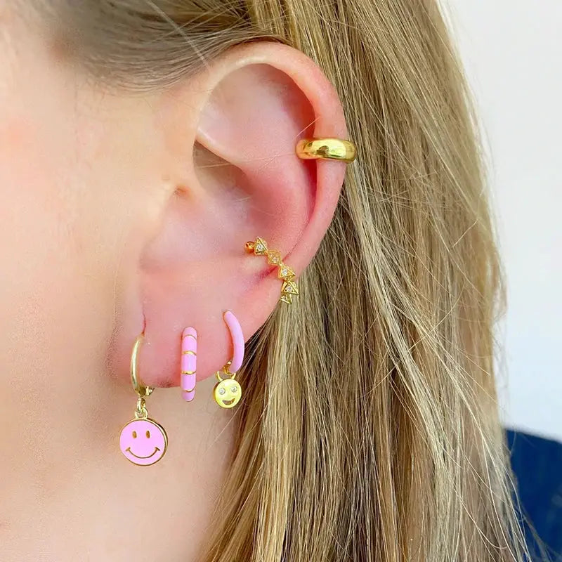 Gold-tone ear jewelry set with pink accents: multiple hoop earrings including smiley pendant and geometric charms worn on the ear